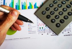 Stock photograph of a person's hand holding an ink pen over a calculator and sheet of paper with bar graphs on it.